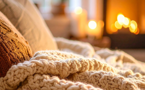 Helle Strickdecke auf Sofa mit Kaminfeuer und Kerzen im Hintergrund